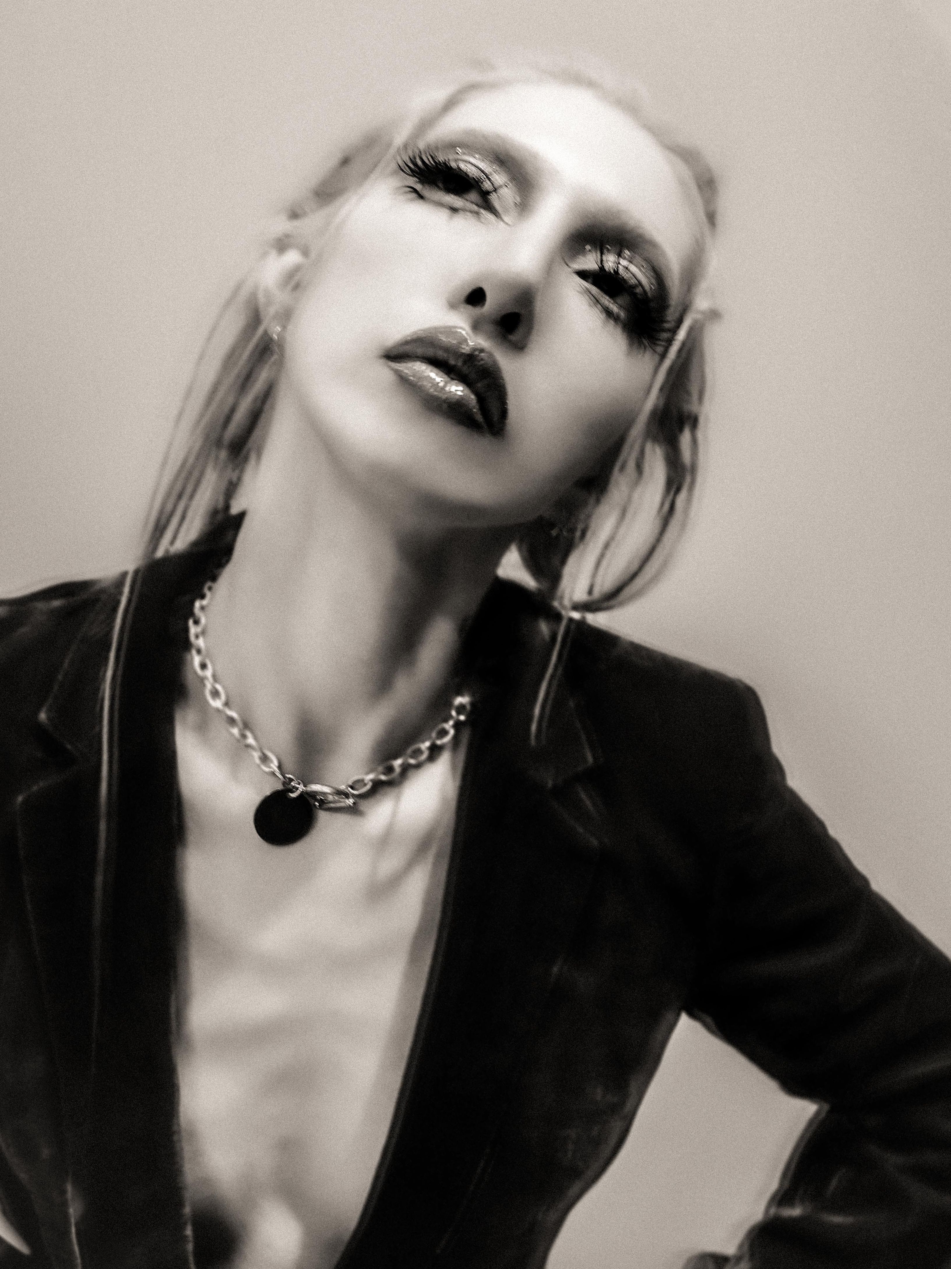 Gemini said A black-and-white, high-angle portrait of a person with long blonde hair, dramatic dark eye makeup, and glossy lips, tilting their head back with a focused expression. They are wearing a dark velvet blazer and a silver chain necklace with a circular pendant against a neutral, grainy background.