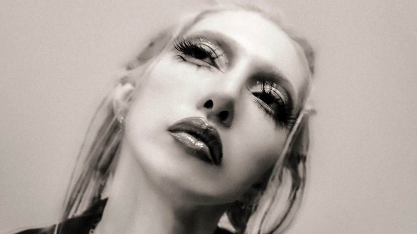 Gemini said A black-and-white, high-angle portrait of a person with long blonde hair, dramatic dark eye makeup, and glossy lips, tilting their head back with a focused expression. They are wearing a dark velvet blazer and a silver chain necklace with a circular pendant against a neutral, grainy background.