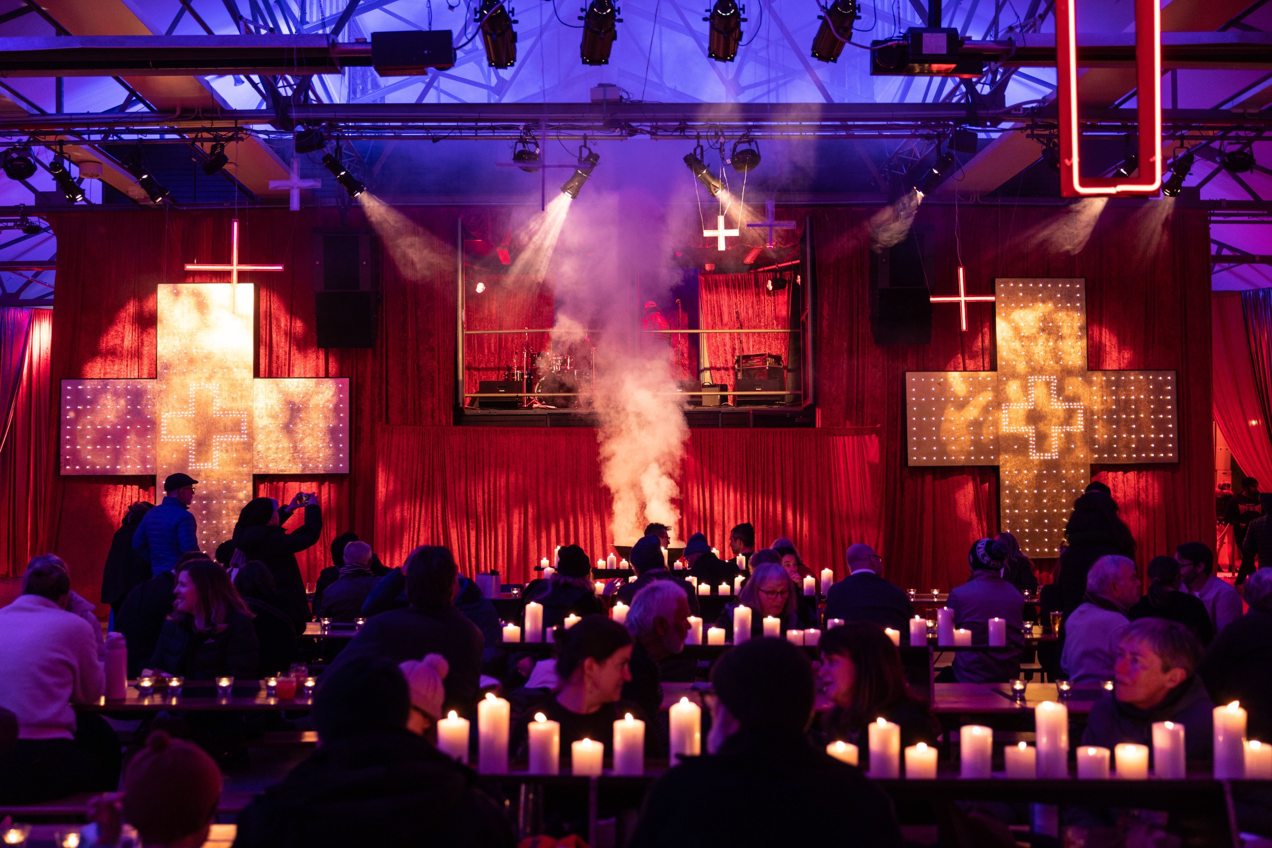 A crowd sits at candlelit tables in a red-lit venue with illuminated crosses on the walls and smoke rising from a central stage.