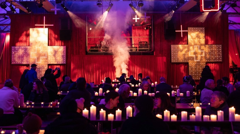 A crowd sits at candlelit tables in a red-lit venue with illuminated crosses on the walls and smoke rising from a central stage.