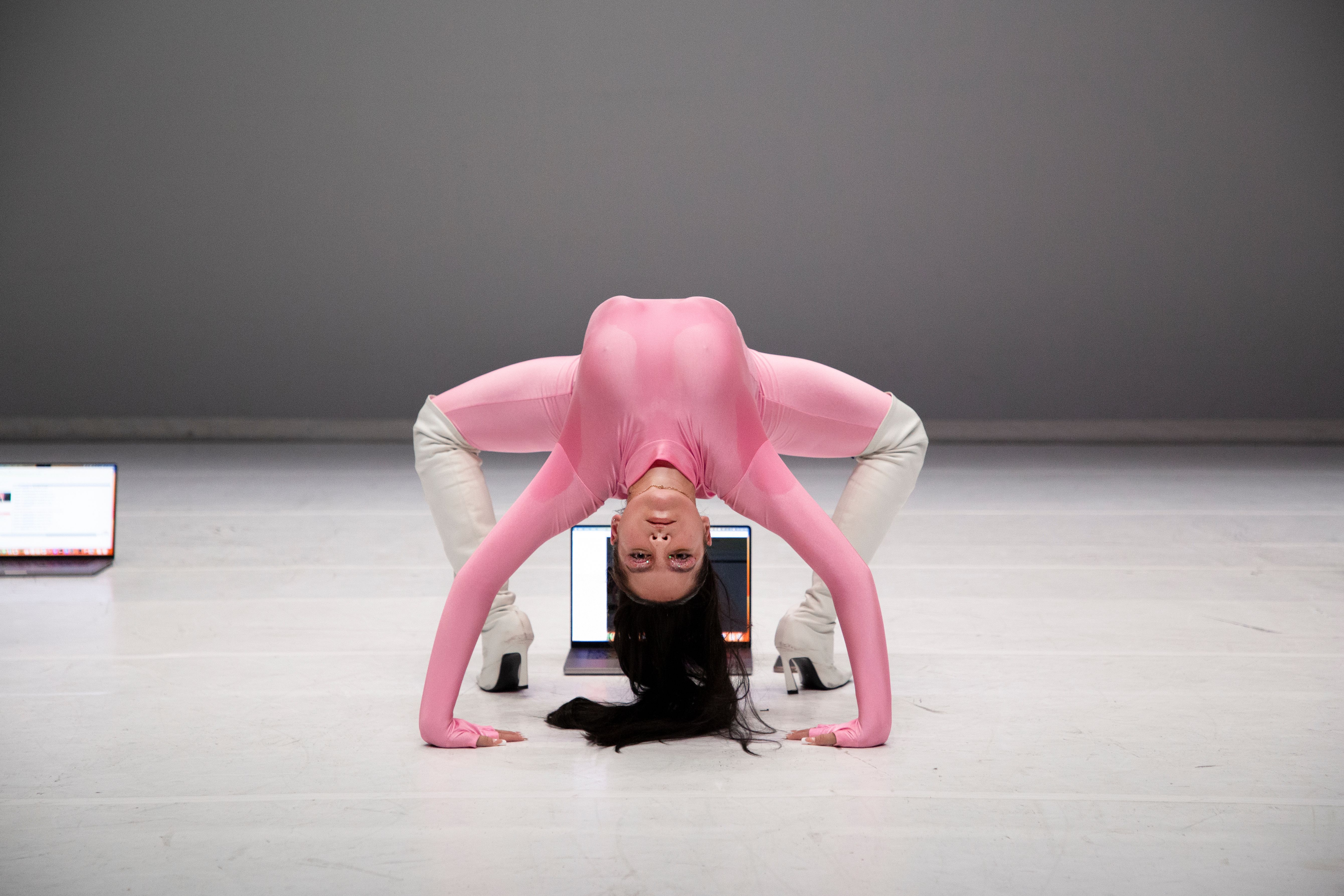 A performer in a pink bodysuit and white boots holds a dramatic, deep backbend on a white stage, looking directly at the camera with their head inverted. Two open laptops are visible on the floor in the background against a minimalist grey wall.