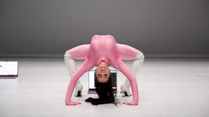 A performer in a pink bodysuit and white boots holds a dramatic, deep backbend on a white stage, looking directly at the camera with their head inverted. Two open laptops are visible on the floor in the background against a minimalist grey wall.