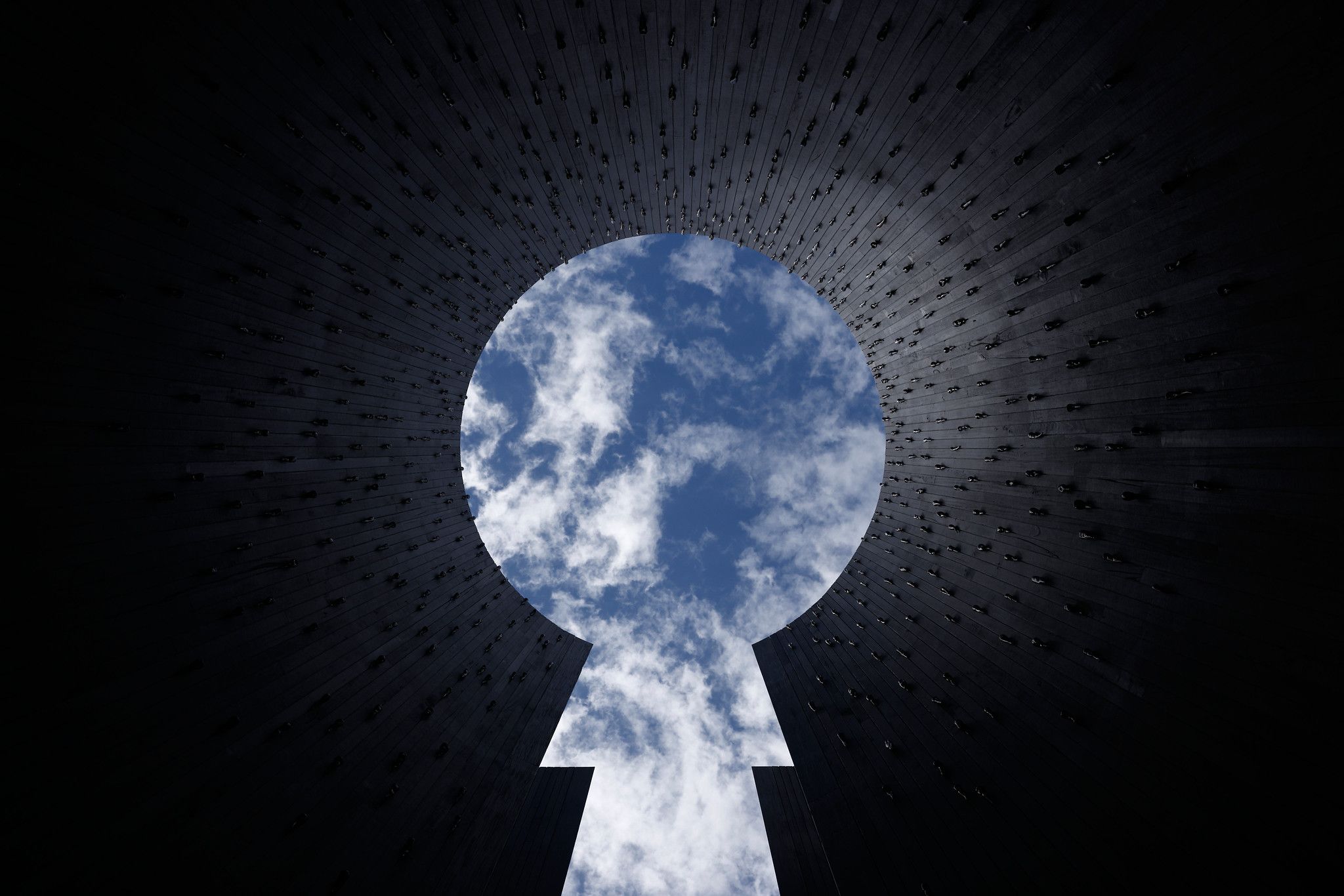 A view looking straight up from inside a tall, dark cylindrical structure with a keyhole-shaped opening at the top. The bright blue sky and white clouds are framed by the silhouette of the walls, which are studded with small metallic objects.