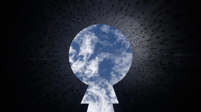 A view looking straight up from inside a tall, dark cylindrical structure with a keyhole-shaped opening at the top. The bright blue sky and white clouds are framed by the silhouette of the walls, which are studded with small metallic objects.