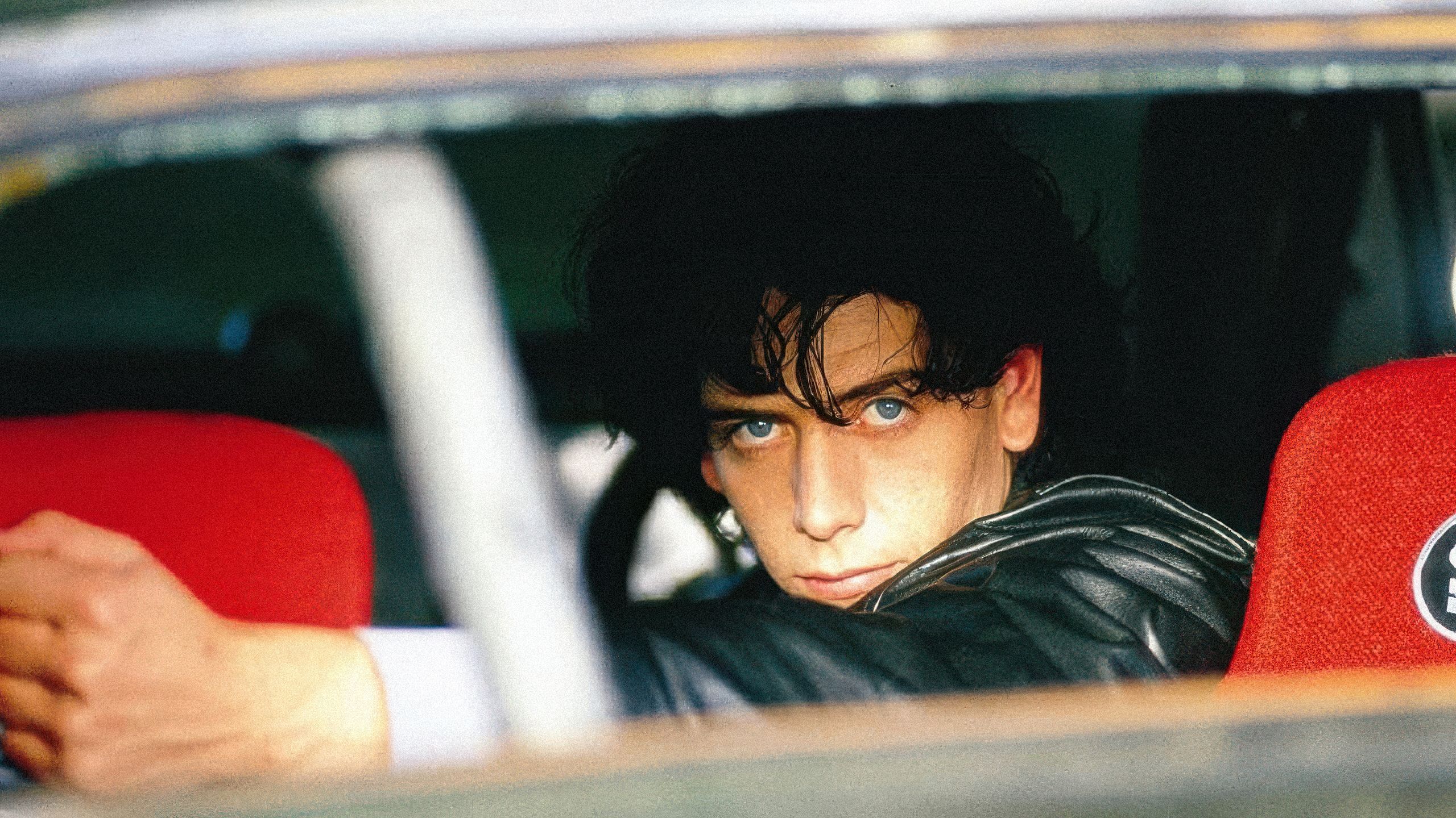 A man with dark, messy hair and striking blue eyes stares intensely from inside a car. The frame is tight, showing his face and one hand on the steering wheel, with a bright red car seat visible behind him.