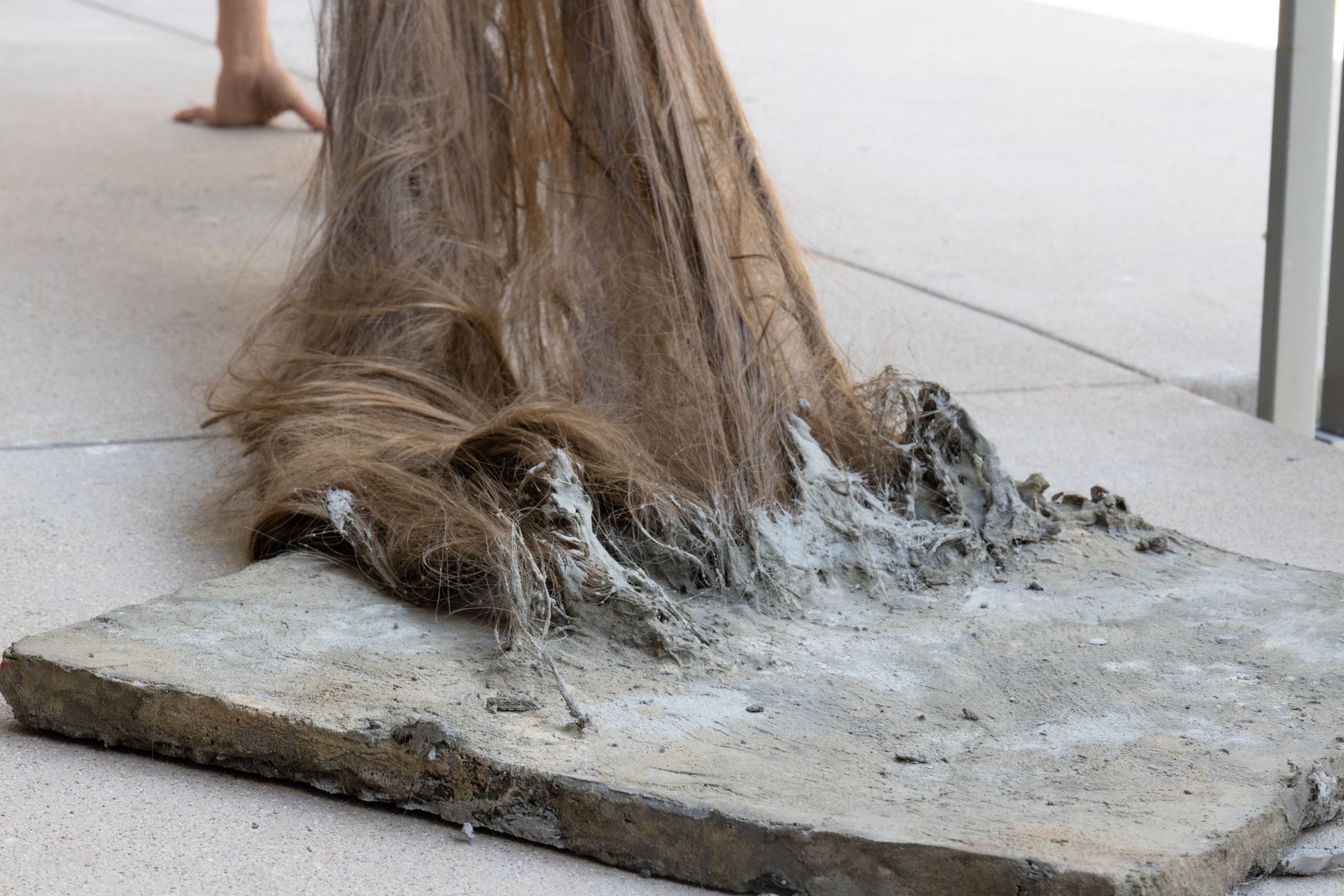 An experimental art piece features a thick mass of long brown hair embedded into a rough concrete slab on the ground. The hair appears to grow out of or merge with the textured grey cement.