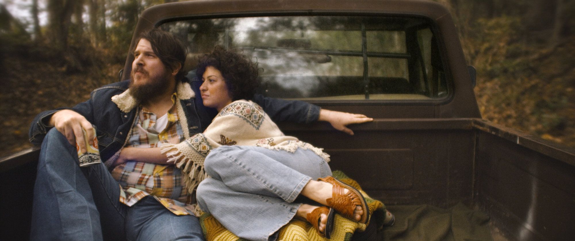 A bearded man in a denim jacket and a woman in a patterned poncho sit close together in the back of a vintage pickup truck. They are parked in a wooded area, surrounded by the soft-focus greenery of a forest.