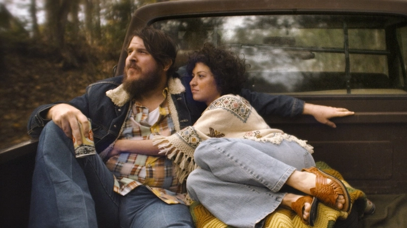 A bearded man in a denim jacket and a woman in a patterned poncho sit close together in the back of a vintage pickup truck. They are parked in a wooded area, surrounded by the soft-focus greenery of a forest.