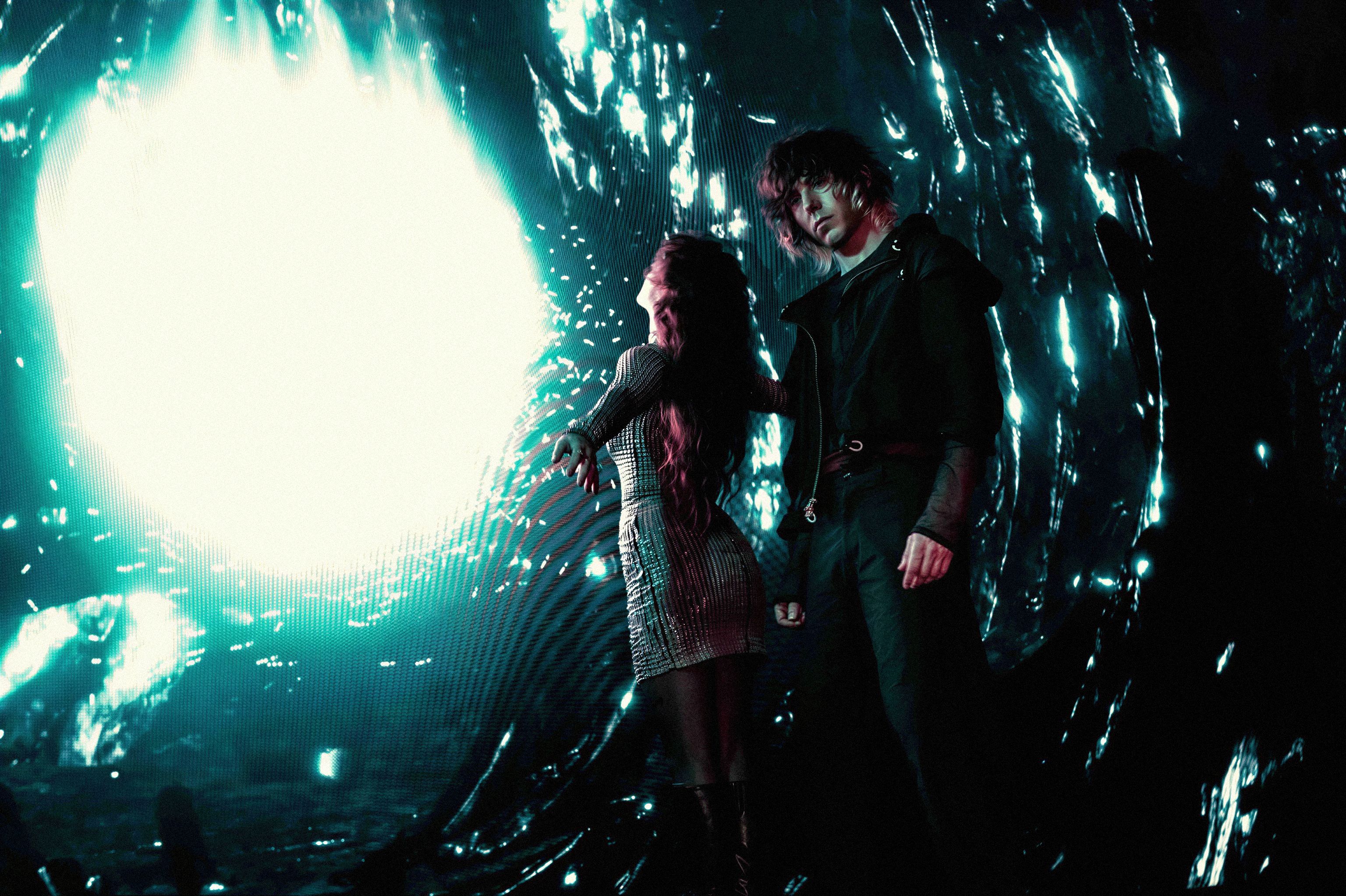 Two figures stand before a massive, glowing teal portal that emits intense light and digital interference patterns. The scene is dark and cinematic, with the silhouettes of the performers framed by the radiant energy.