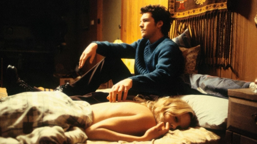 A man in a blue sweater sits on the edge of a bed, looking off-camera, while a blonde woman lies face down in the foreground. The room is dimly lit with warm wood paneling, suggesting a quiet, somber moment.