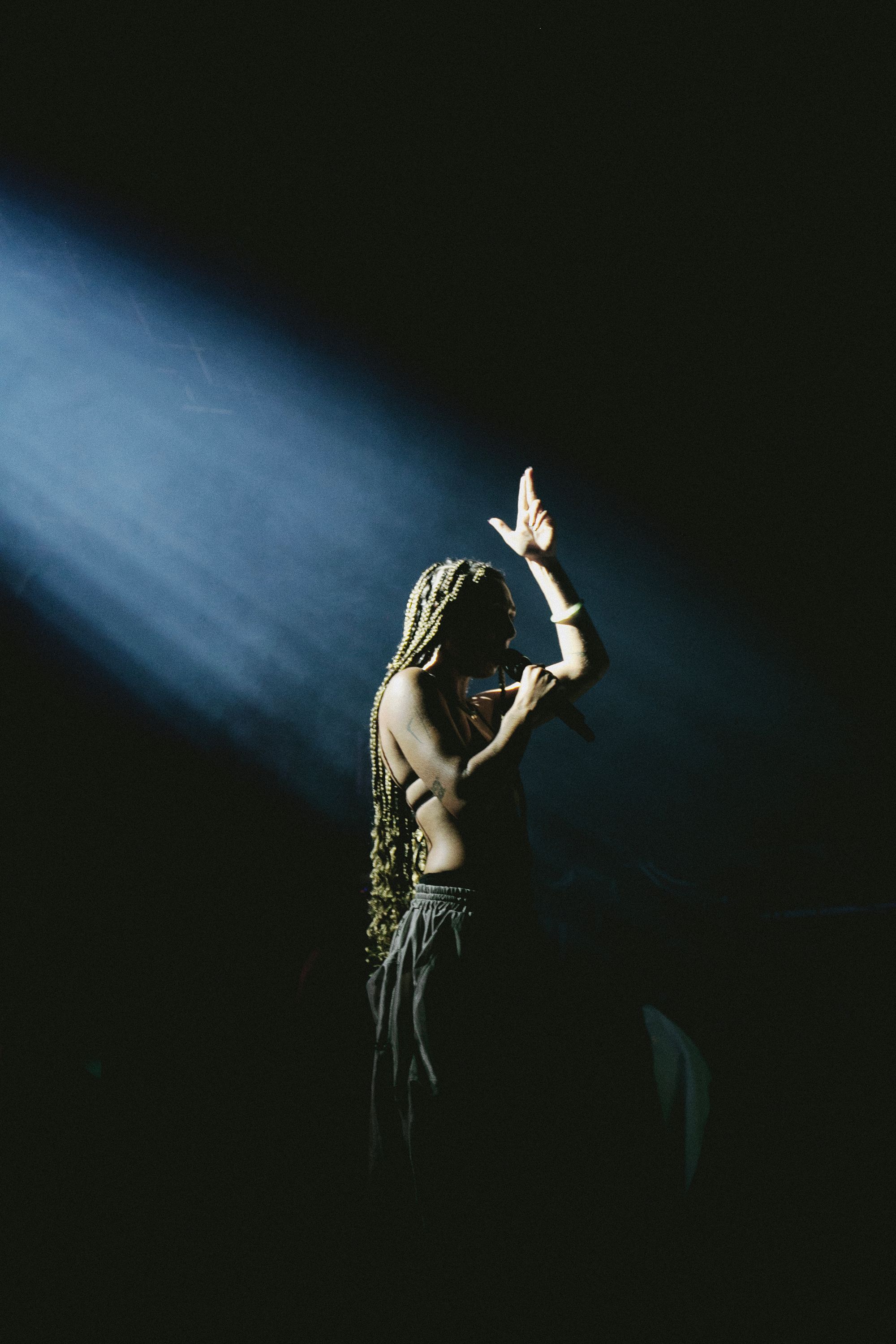 A dramatic, high-contrast shot of a performer with long braids standing in a single bright spotlight against a pitch-black background. She holds a microphone to her lips and raises one hand upward, capturing an intense, powerful moment on stage.