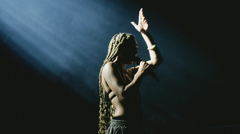 A dramatic, high-contrast shot of a performer with long braids standing in a single bright spotlight against a pitch-black background. She holds a microphone to her lips and raises one hand upward, capturing an intense, powerful moment on stage.