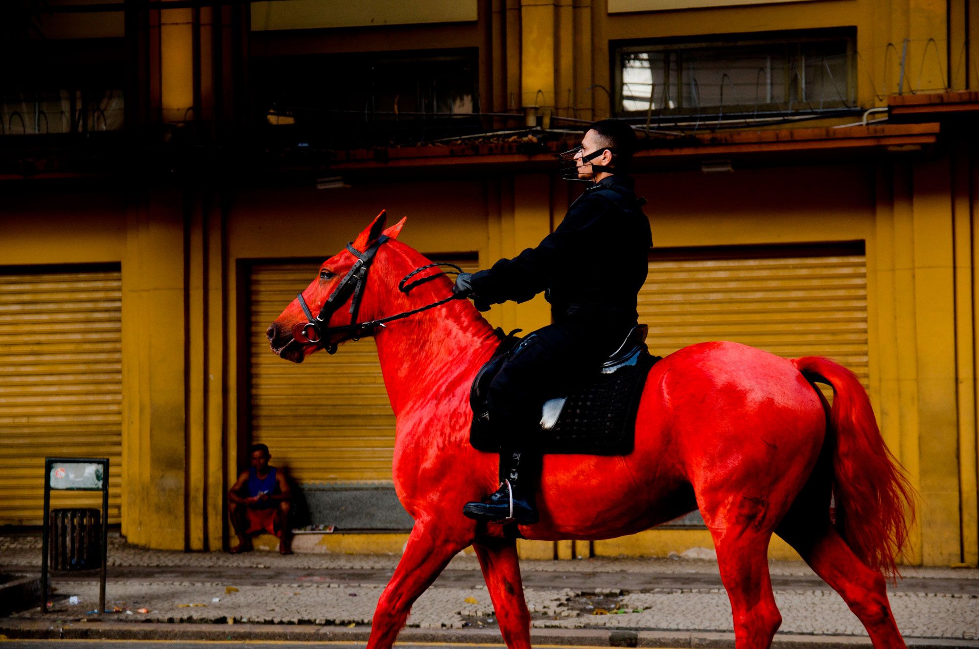 A person in a black uniform and a muzzle-like mask rides a vibrant, bright red horse past a mustard-yellow building. A person sits on the sidewalk in the background.