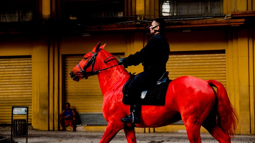A person in a black uniform and a muzzle-like mask rides a vibrant, bright red horse past a mustard-yellow building. A person sits on the sidewalk in the background.