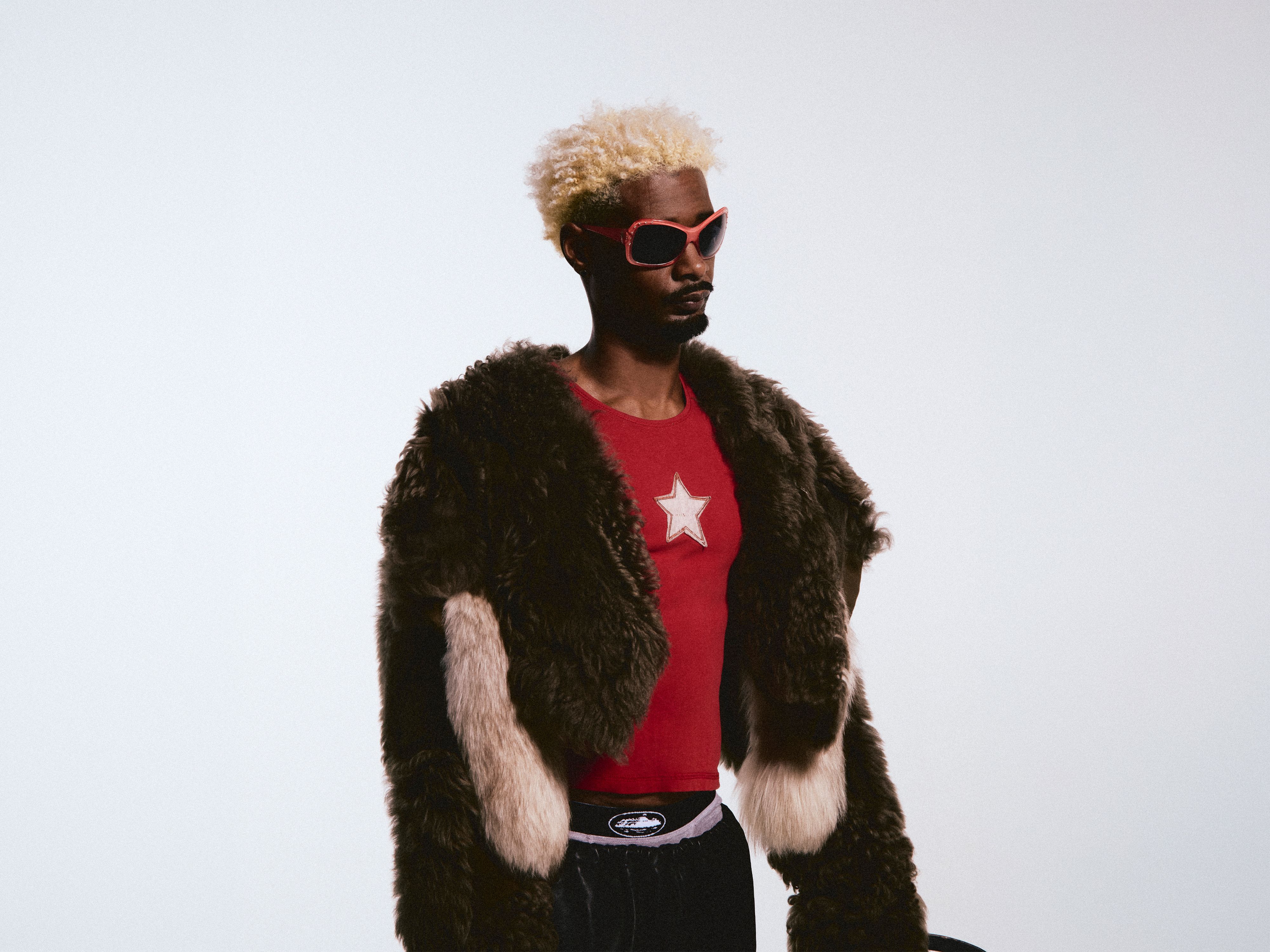 A man with bleached blonde hair and red-rimmed sunglasses poses in a high-fashion outfit against a white background. He wears a red shirt with a white star emblem underneath a bulky, multi-toned faux fur coat.