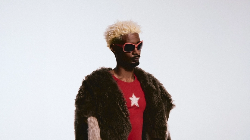 A man with bleached blonde hair and red-rimmed sunglasses poses in a high-fashion outfit against a white background. He wears a red shirt with a white star emblem underneath a bulky, multi-toned faux fur coat.