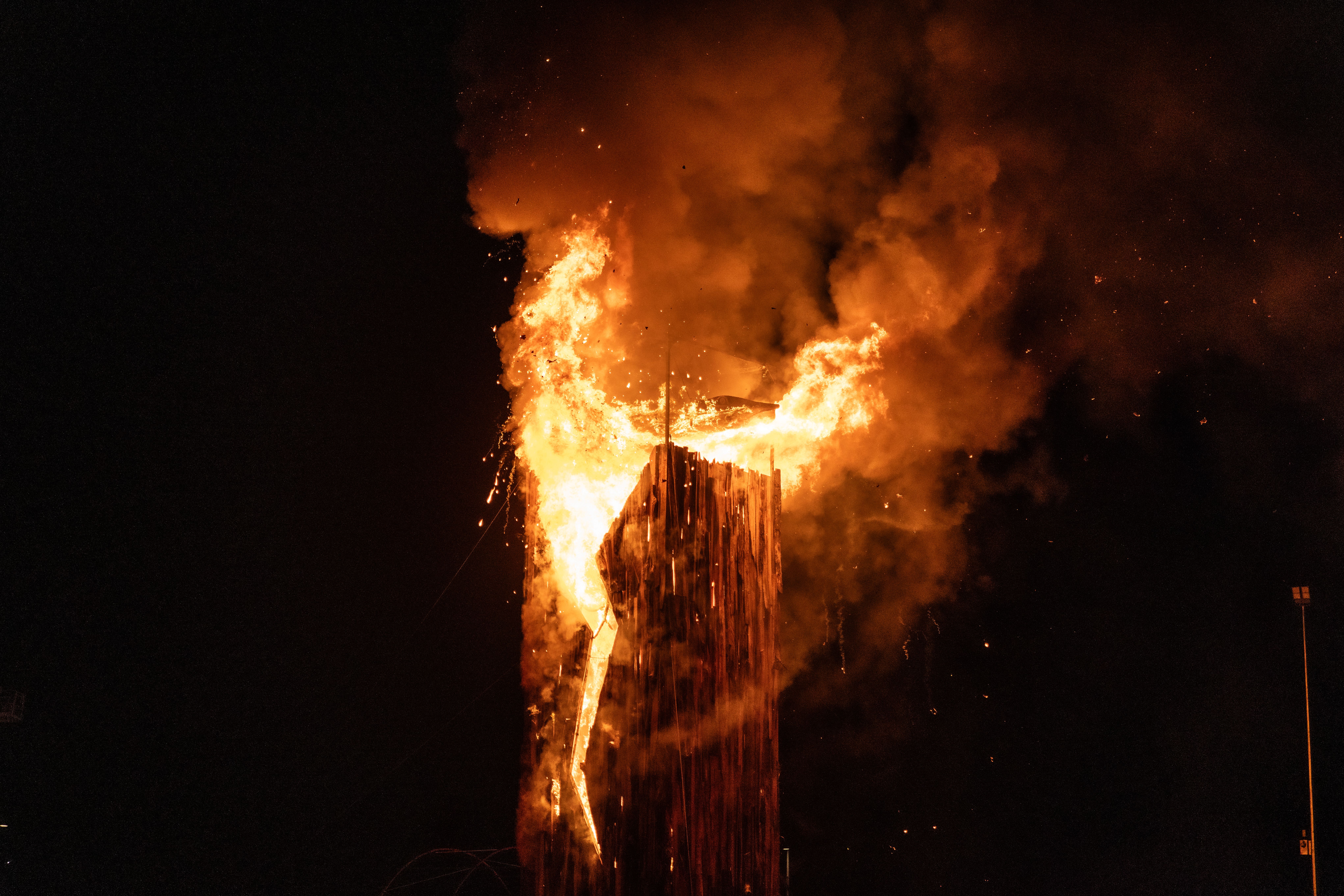 A tall wooden structure is engulfed in massive orange flames and thick smoke against a pitch-black night sky, with sparks falling.