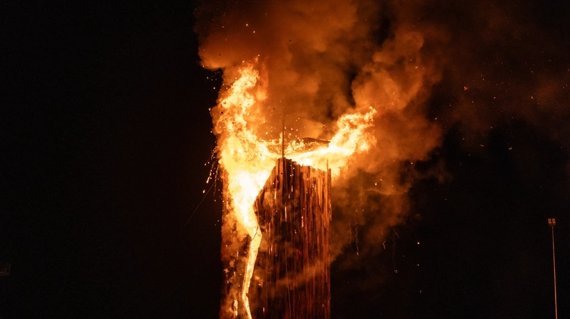 A tall wooden structure is engulfed in massive orange flames and thick smoke against a pitch-black night sky, with sparks falling.
