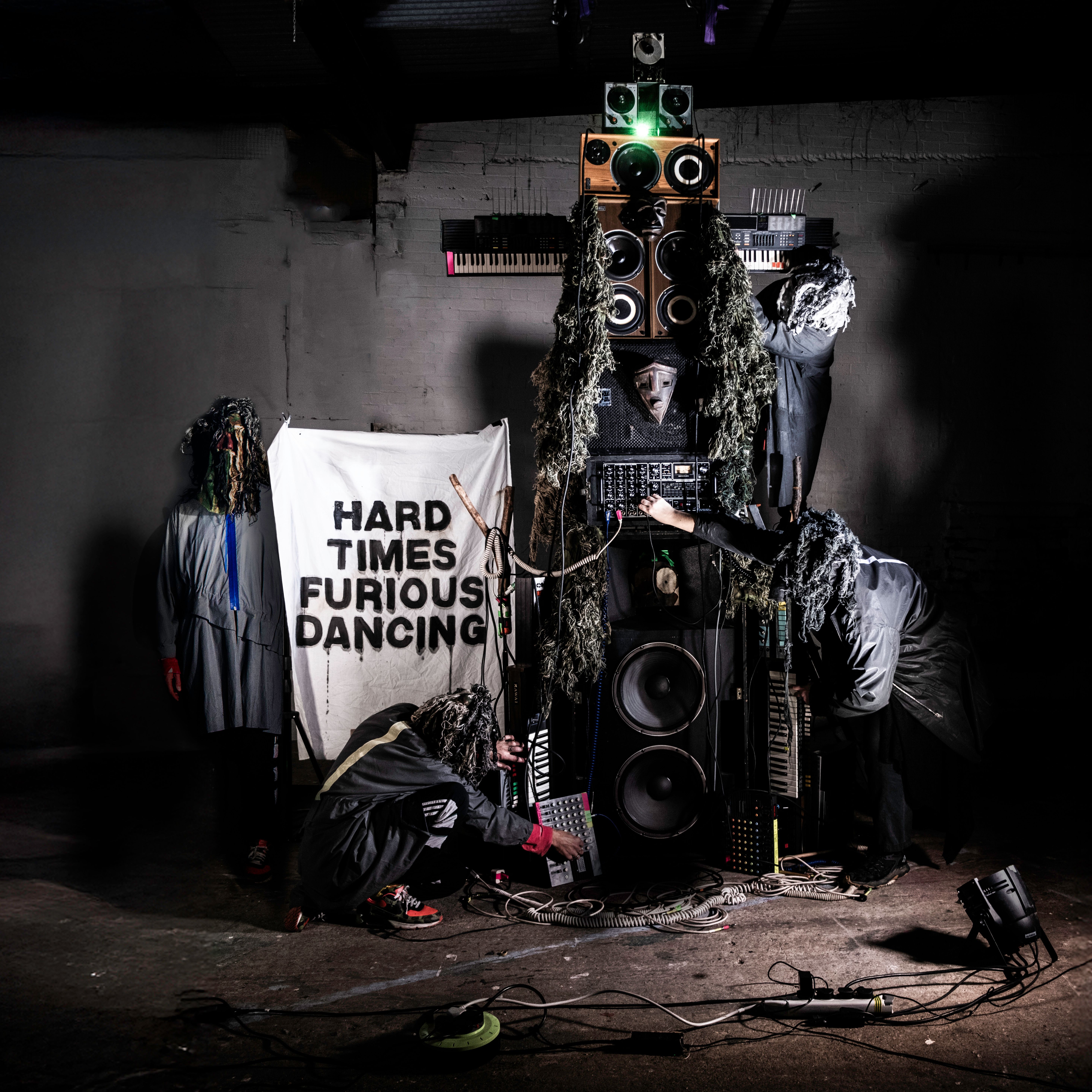 A band in shaggy, forest-like costumes performs around a DIY totem pole of speakers and synthesizers. A banner in the background reads "HARD TIMES FURIOUS DANCING" in bold, black block letters.