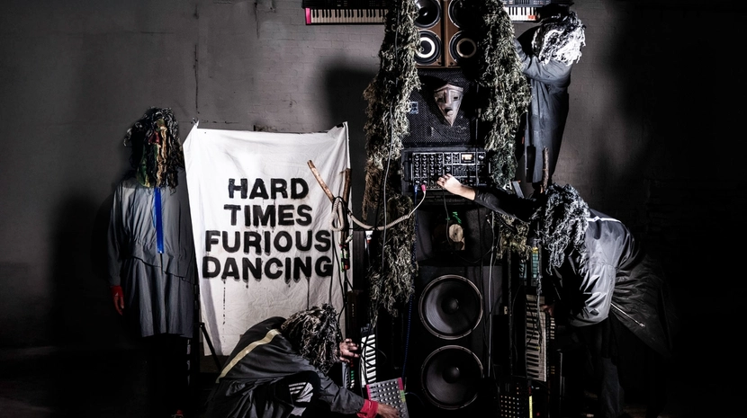 A band in shaggy, forest-like costumes performs around a DIY totem pole of speakers and synthesizers. A banner in the background reads "HARD TIMES FURIOUS DANCING" in bold, black block letters.