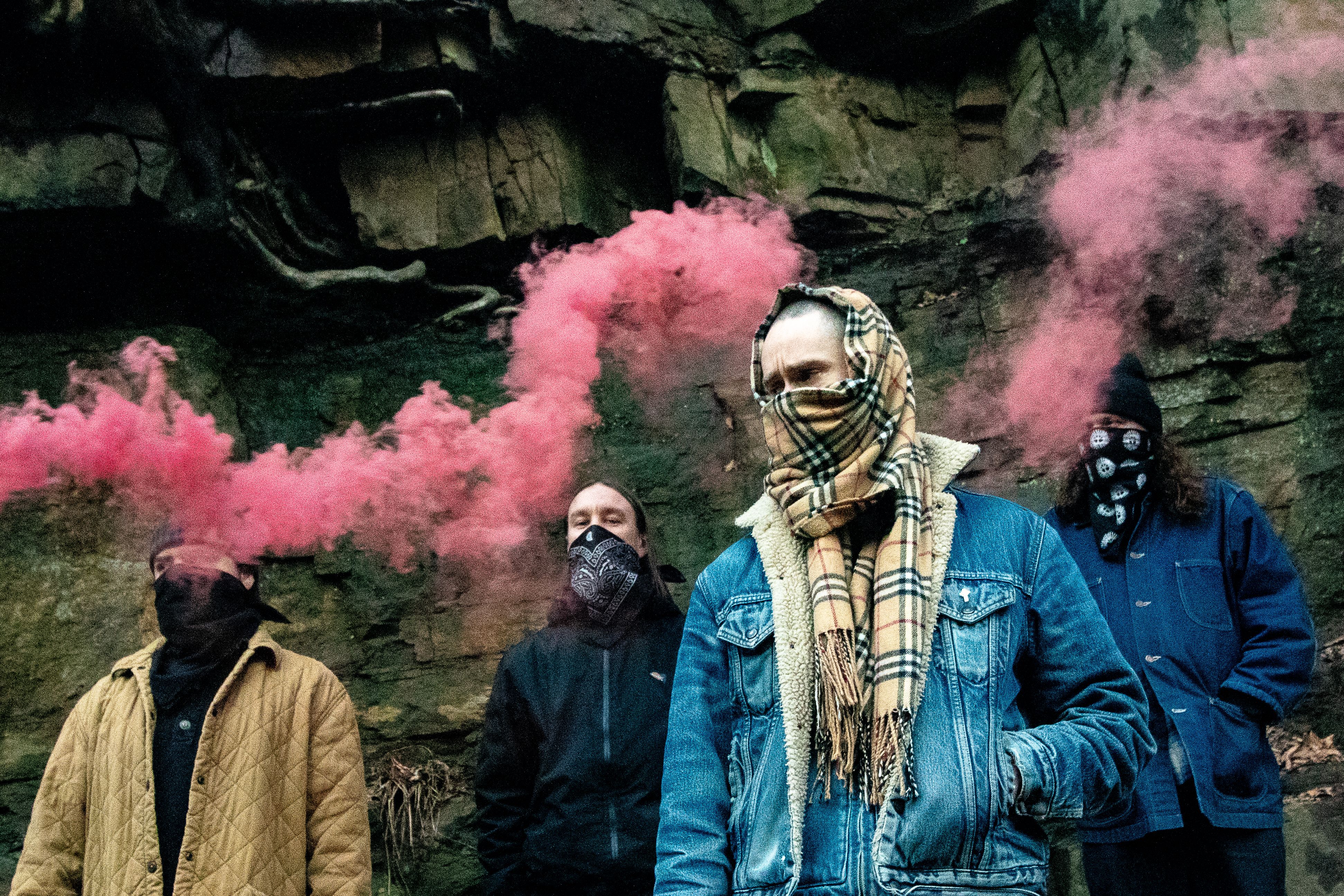 Four people with their faces obscured by scarves and bandanas stand in front of a rocky cliff side. Vibrant pink smoke plumes drift across the scene as they pose in casual jackets and winter gear.
