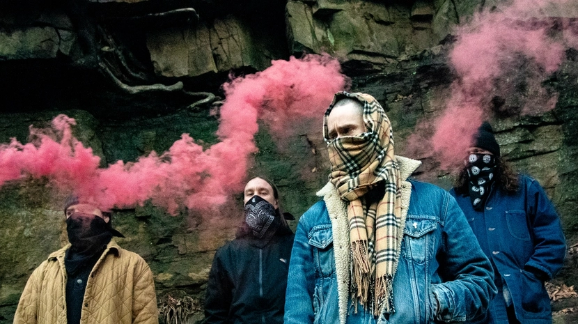 Four people with their faces obscured by scarves and bandanas stand in front of a rocky cliff side. Vibrant pink smoke plumes drift across the scene as they pose in casual jackets and winter gear.