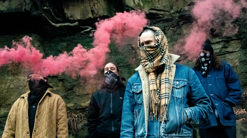 Four people with their faces obscured by scarves and bandanas stand in front of a rocky cliff side. Vibrant pink smoke plumes drift across the scene as they pose in casual jackets and winter gear.