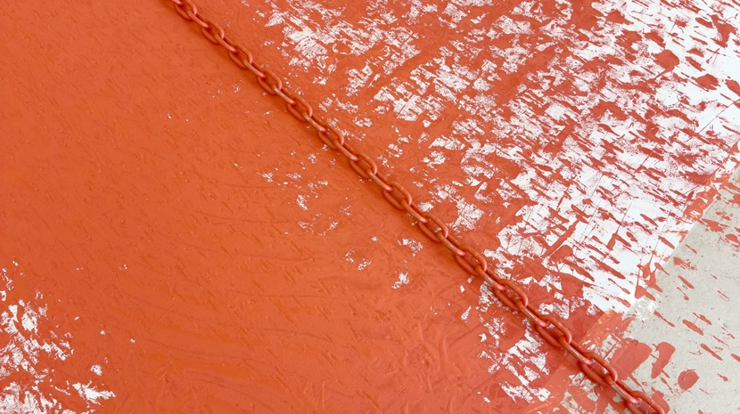 A orange-and-white, high-angle shot shows a thick metal chain stretched horizontally across a floor with contrasting textures. The left side of the floor is covered in a dark, glossy, and textured substance resembling wet paint or tar, while the right side is a light, matte, and uniform surface.