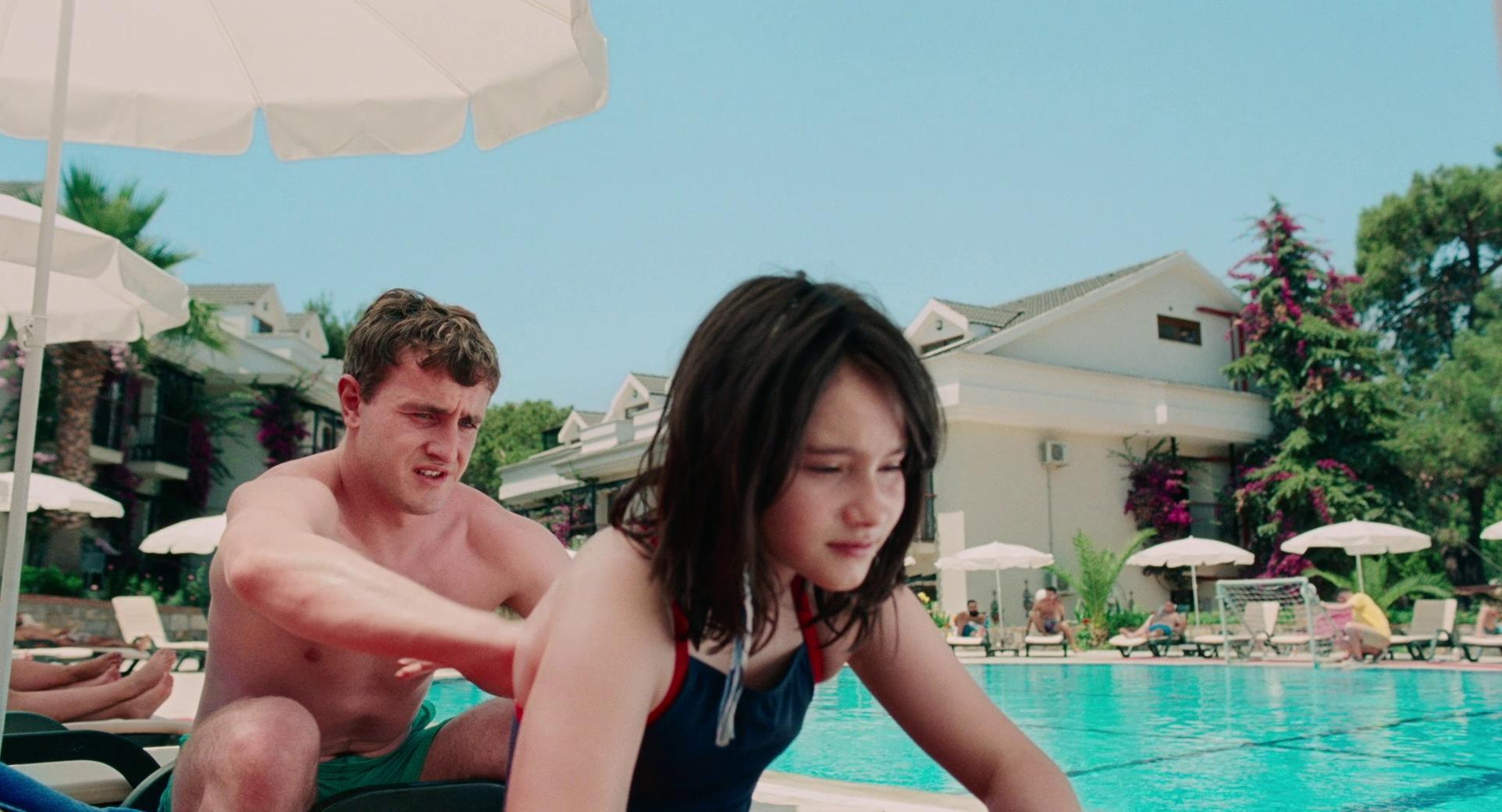 A sun-drenched scene at a holiday resort where a man in green swim trunks applies sunscreen to a young girl's back by a bright blue pool. The background features white patio umbrellas and Mediterranean-style villas under a clear, pale sky.