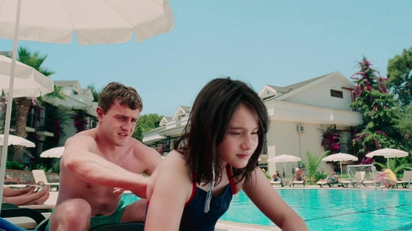 A sun-drenched scene at a holiday resort where a man in green swim trunks applies sunscreen to a young girl's back by a bright blue pool. The background features white patio umbrellas and Mediterranean-style villas under a clear, pale sky.