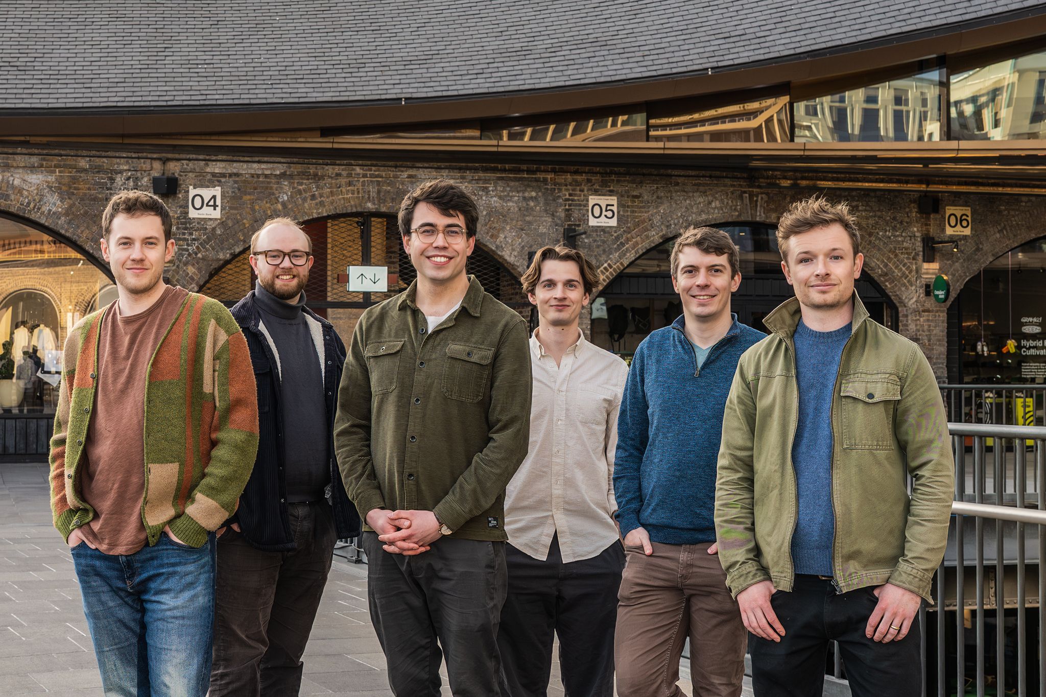 Six men in casual contemporary clothing stand in a line, smiling for a group portrait in an outdoor urban space. The background features brick arches and a modern curved roofline under bright, even daylight.