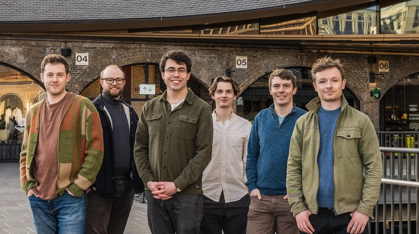 Six men in casual contemporary clothing stand in a line, smiling for a group portrait in an outdoor urban space. The background features brick arches and a modern curved roofline under bright, even daylight.