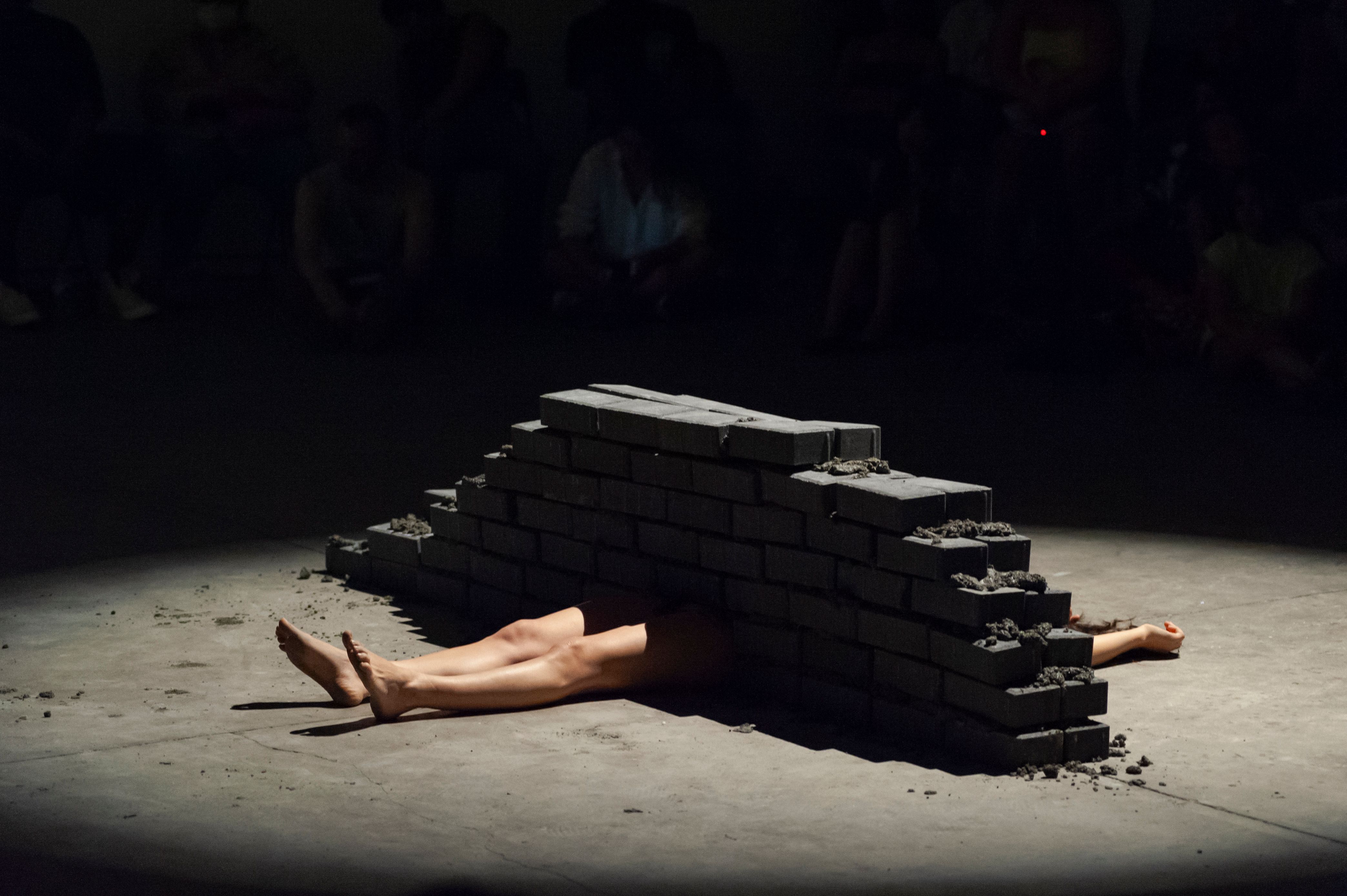 A person lies flat on the floor with their torso completely obscured by a low, jagged wall built of dark bricks. A single spotlight illuminates the scene, highlighting the bare legs and one arm extending from either side of the barrier.