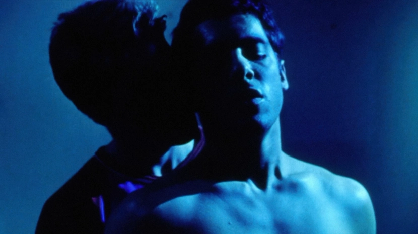 A tight, intimate shot of two men's heads in profile, bathed in a deep, moody blue light. The lighting creates heavy shadows, emphasizing the emotional tension and physical closeness between the figures.