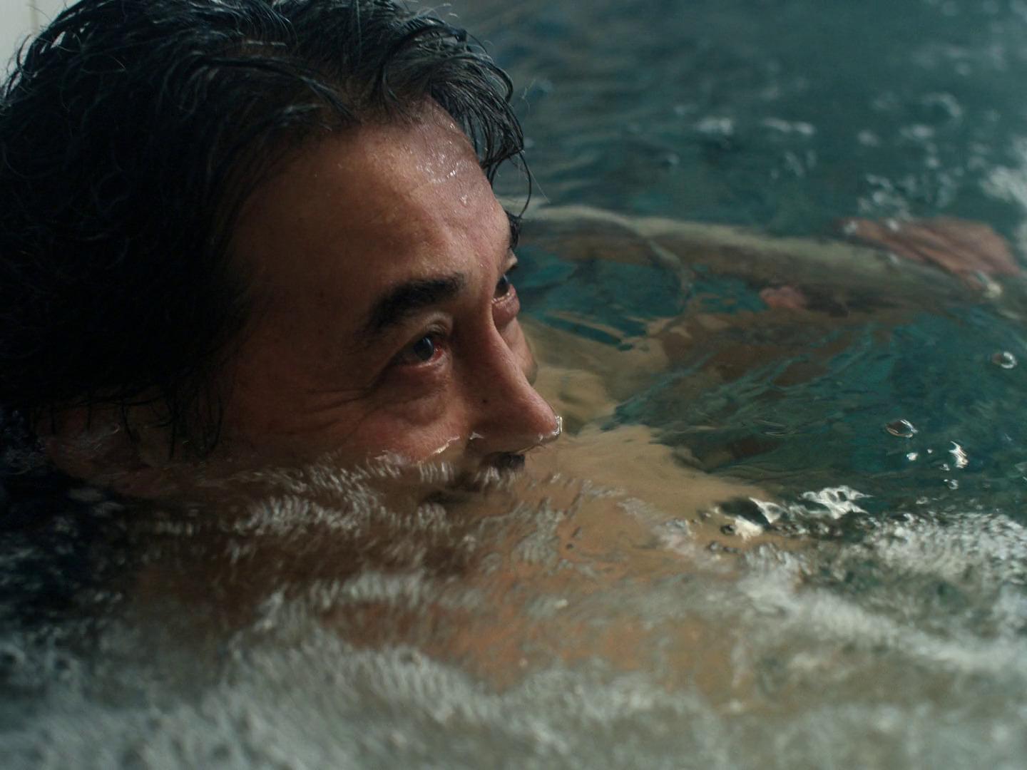 A close-up, low-angle shot of a man’s face as he soaks in water, with only his eyes and nose above the surface. Reflections and ripples in the water create a serene, contemplative atmosphere.
