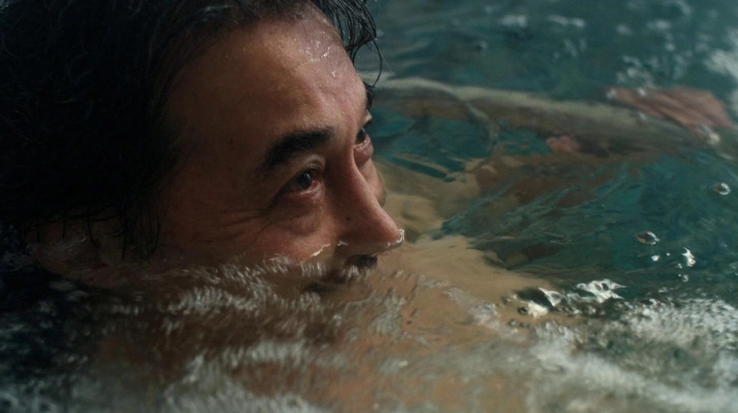 A close-up, low-angle shot of a man’s face as he soaks in water, with only his eyes and nose above the surface. Reflections and ripples in the water create a serene, contemplative atmosphere.