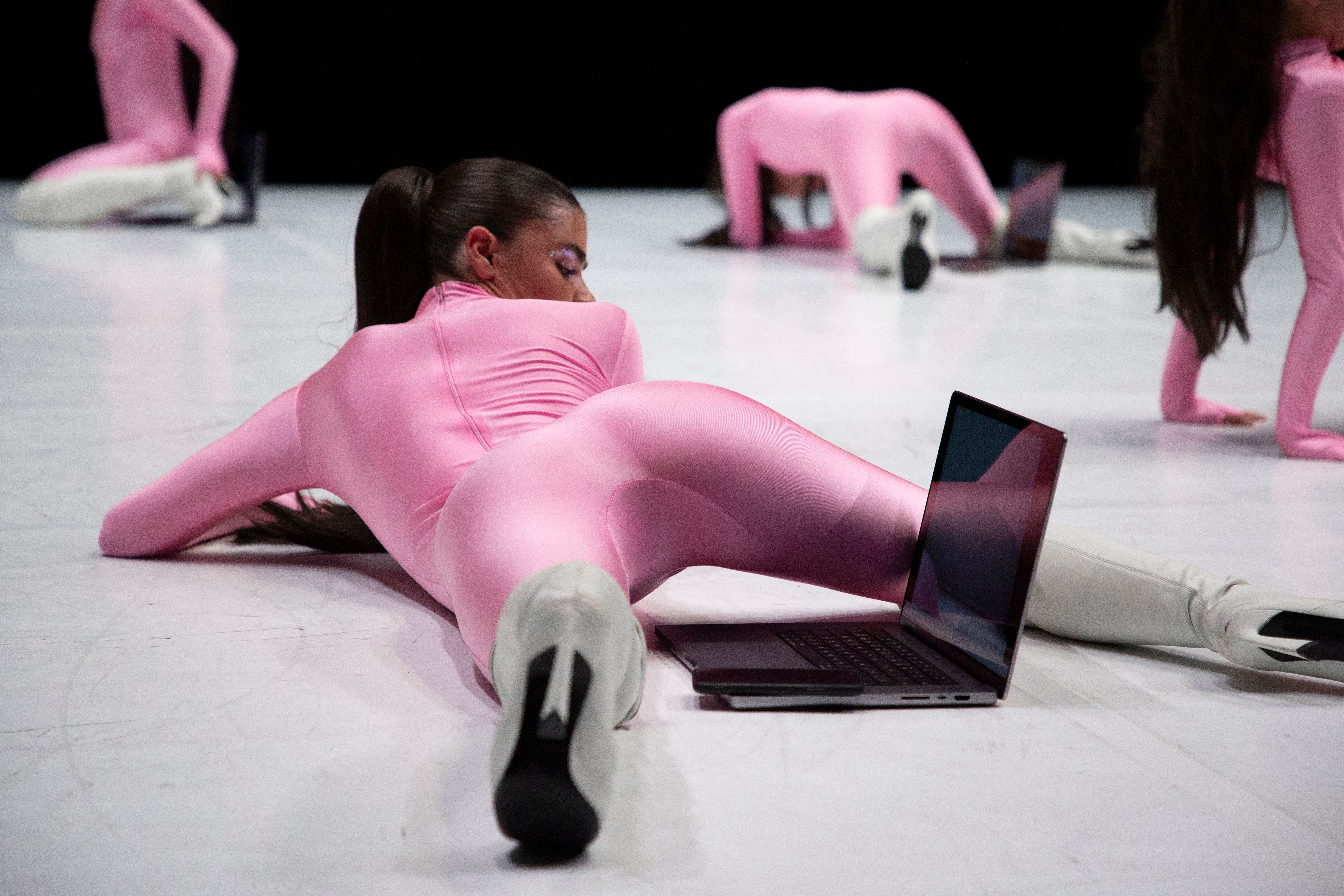 Dancers in sleek, bubblegum pink bodysuits and white boots perform on a bright white floor. One dancer in the foreground lies on her stomach, looking back toward a laptop open on the floor next to her.