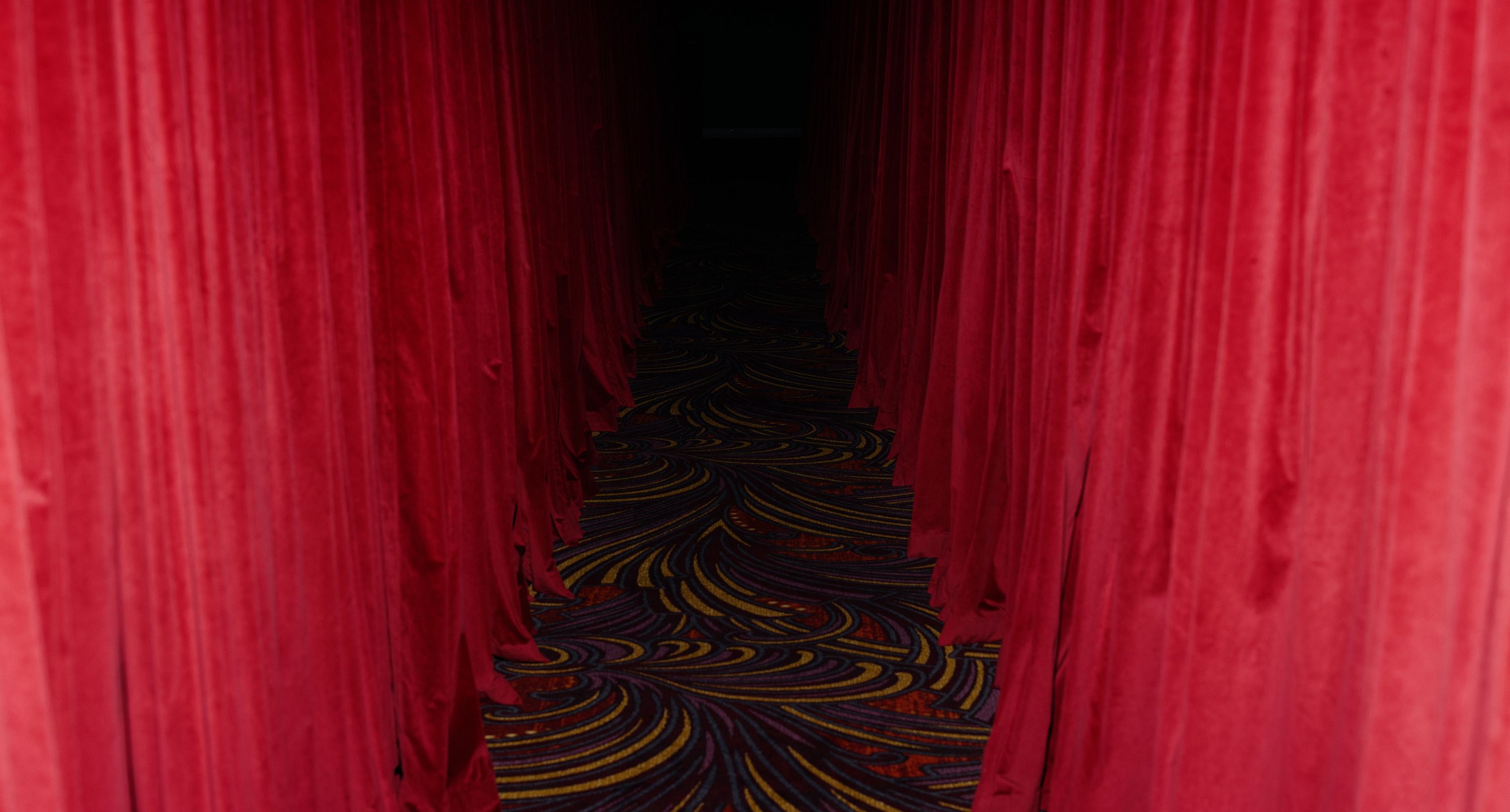A perspective shot looking down a narrow, dimly lit corridor lined with heavy red velvet curtains. The floor is covered in a patterned carpet with swirling gold and purple designs that lead toward a dark, shrouded doorway.