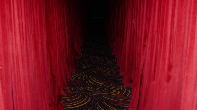 A perspective shot looking down a narrow, dimly lit corridor lined with heavy red velvet curtains. The floor is covered in a patterned carpet with swirling gold and purple designs that lead toward a dark, shrouded doorway.