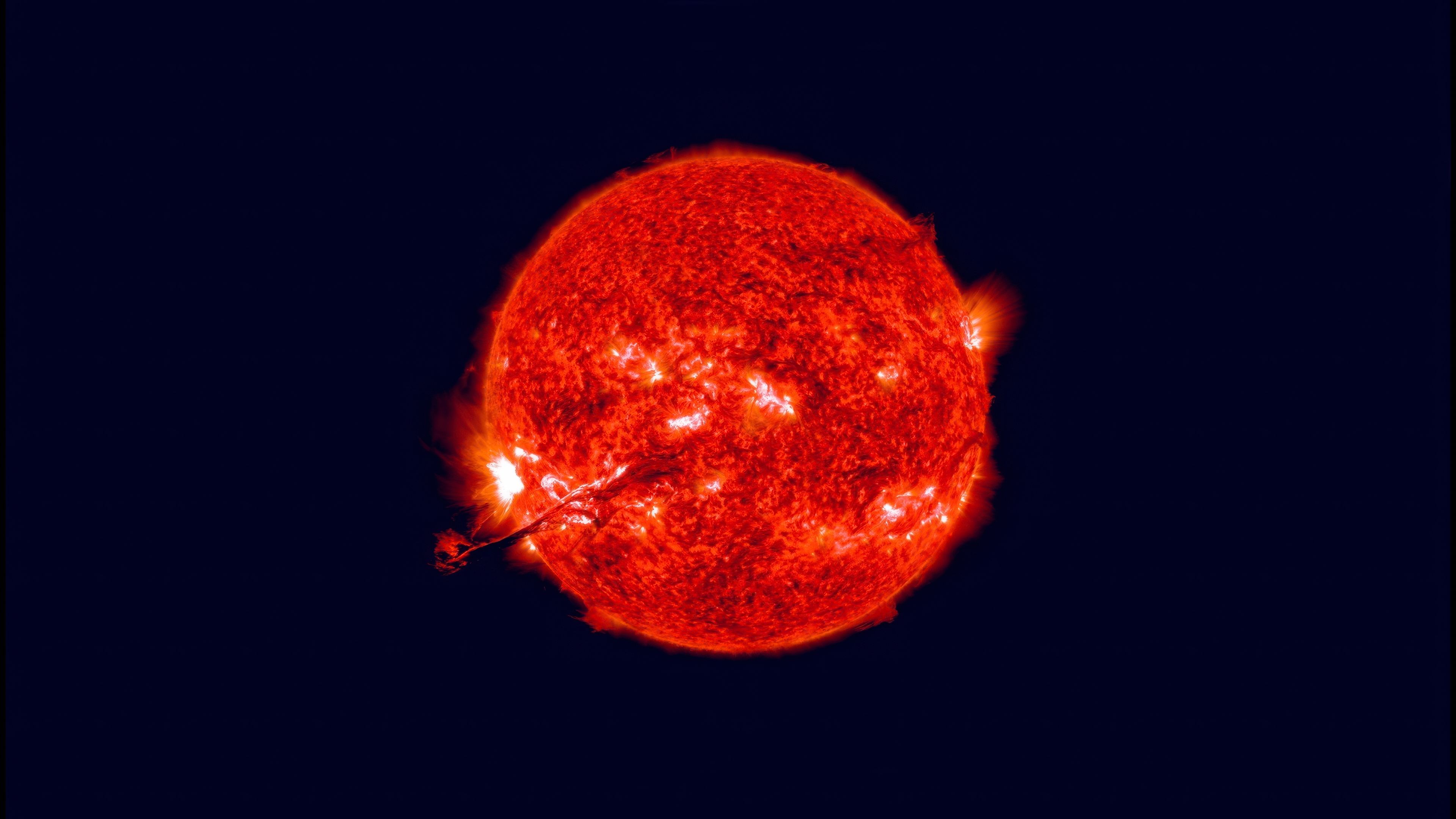 A high-contrast, glowing red sun with visible solar flares and surface textures against a pitch-black background. A prominent solar prominence erupts from the lower left side of the star.