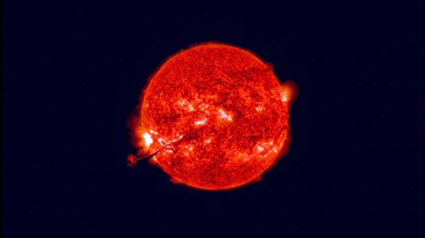 A high-contrast, glowing red sun with visible solar flares and surface textures against a pitch-black background. A prominent solar prominence erupts from the lower left side of the star.