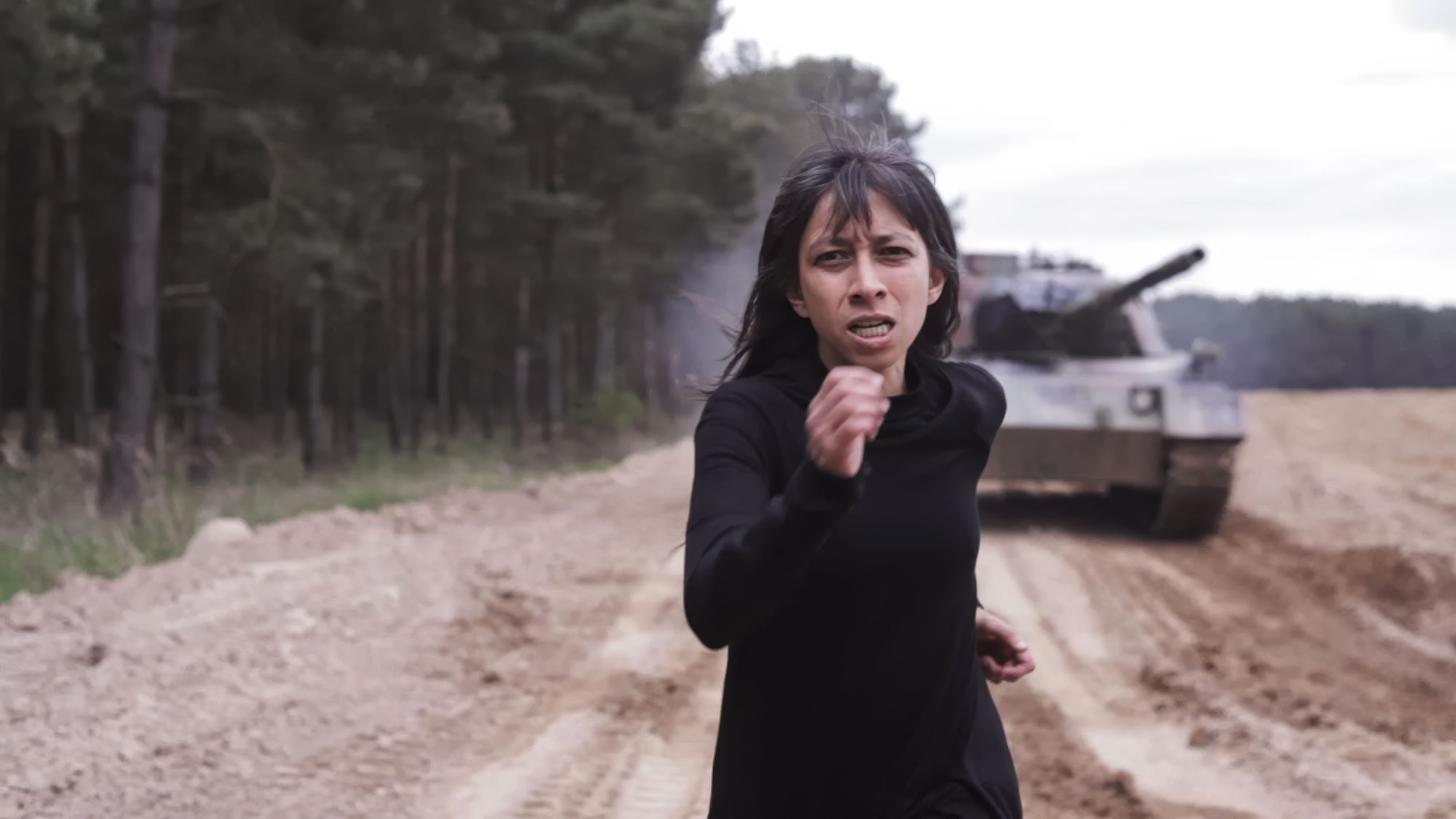 A woman in black clothing runs down a dirt path with a determined, strained expression as a large military tank pursues her from behind. The scene is set in a desolate, wooded landscape under an overcast sky.
