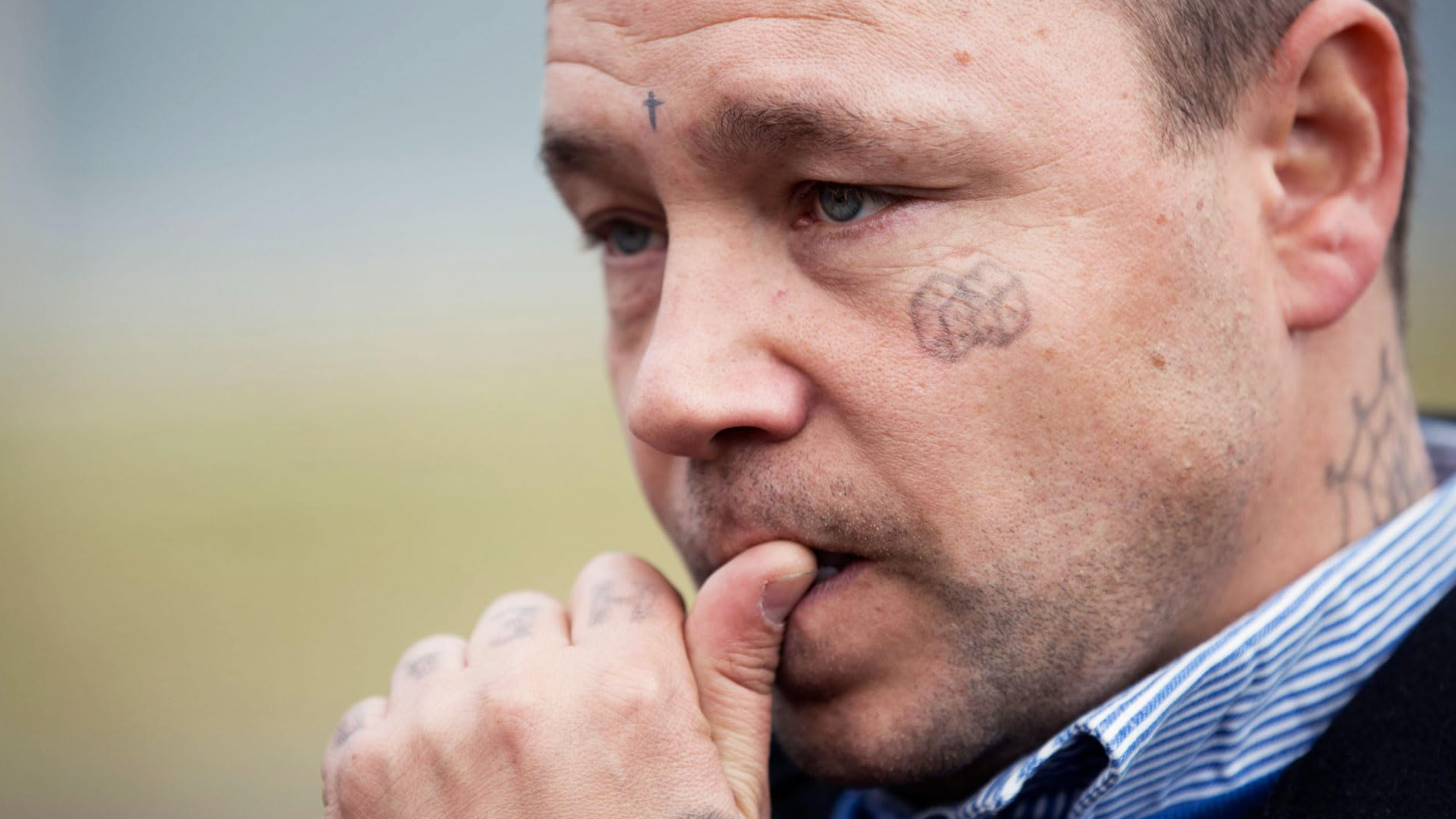 A tight close-up of a man with a troubled expression, biting his thumb in thought. He has several small tattoos on his face and neck, including a cross on his forehead and a spiderweb behind his ear, set against a blurred outdoor background.