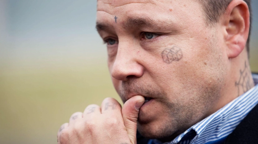 A tight close-up of a man with a troubled expression, biting his thumb in thought. He has several small tattoos on his face and neck, including a cross on his forehead and a spiderweb behind his ear, set against a blurred outdoor background.