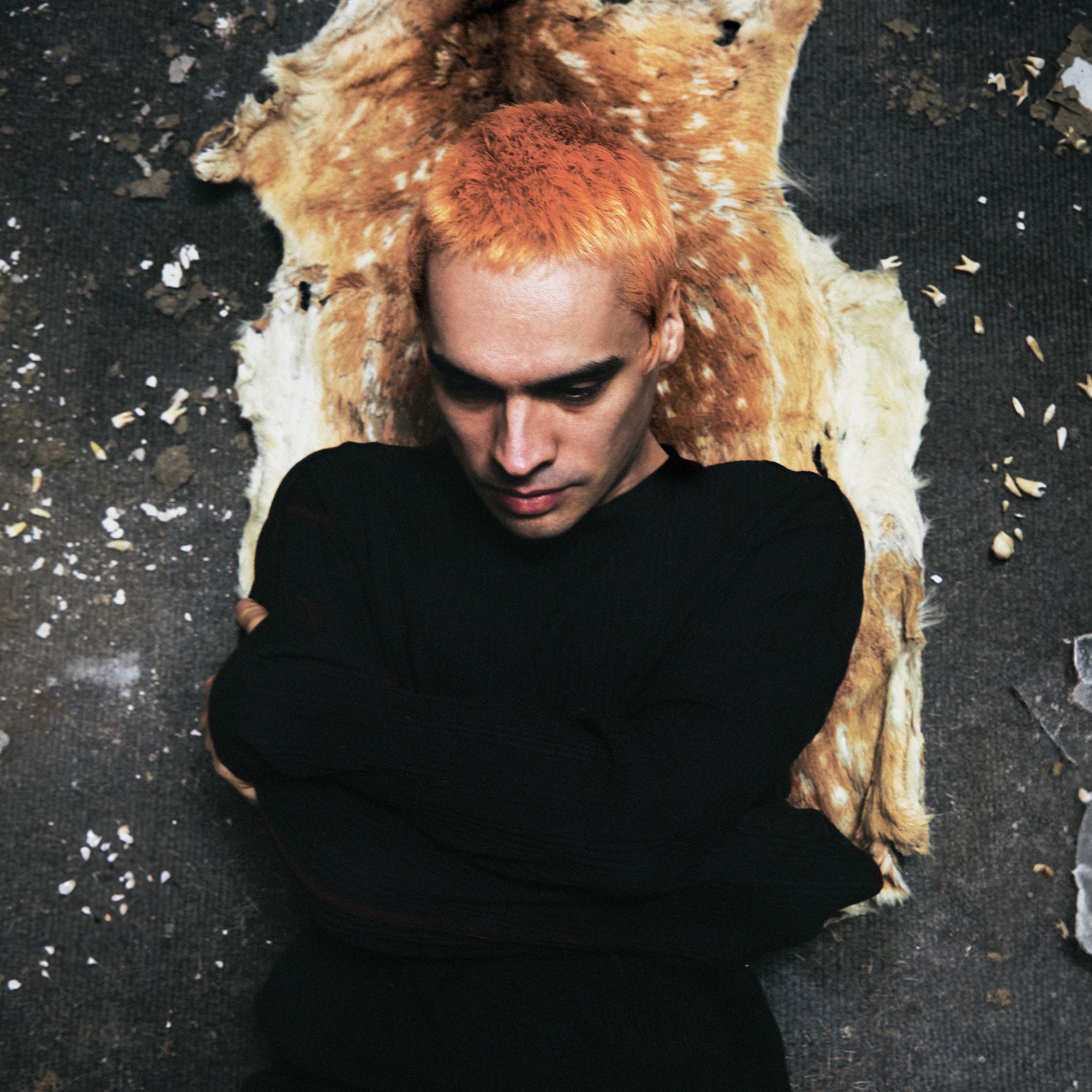 Gemini said A man with short, bright orange hair lies on a dark, textured floor, resting his head on a spotted animal pelt while hugging himself in a black long-sleeved shirt. Small white debris and what appear to be scattered teeth surround him on the ground.