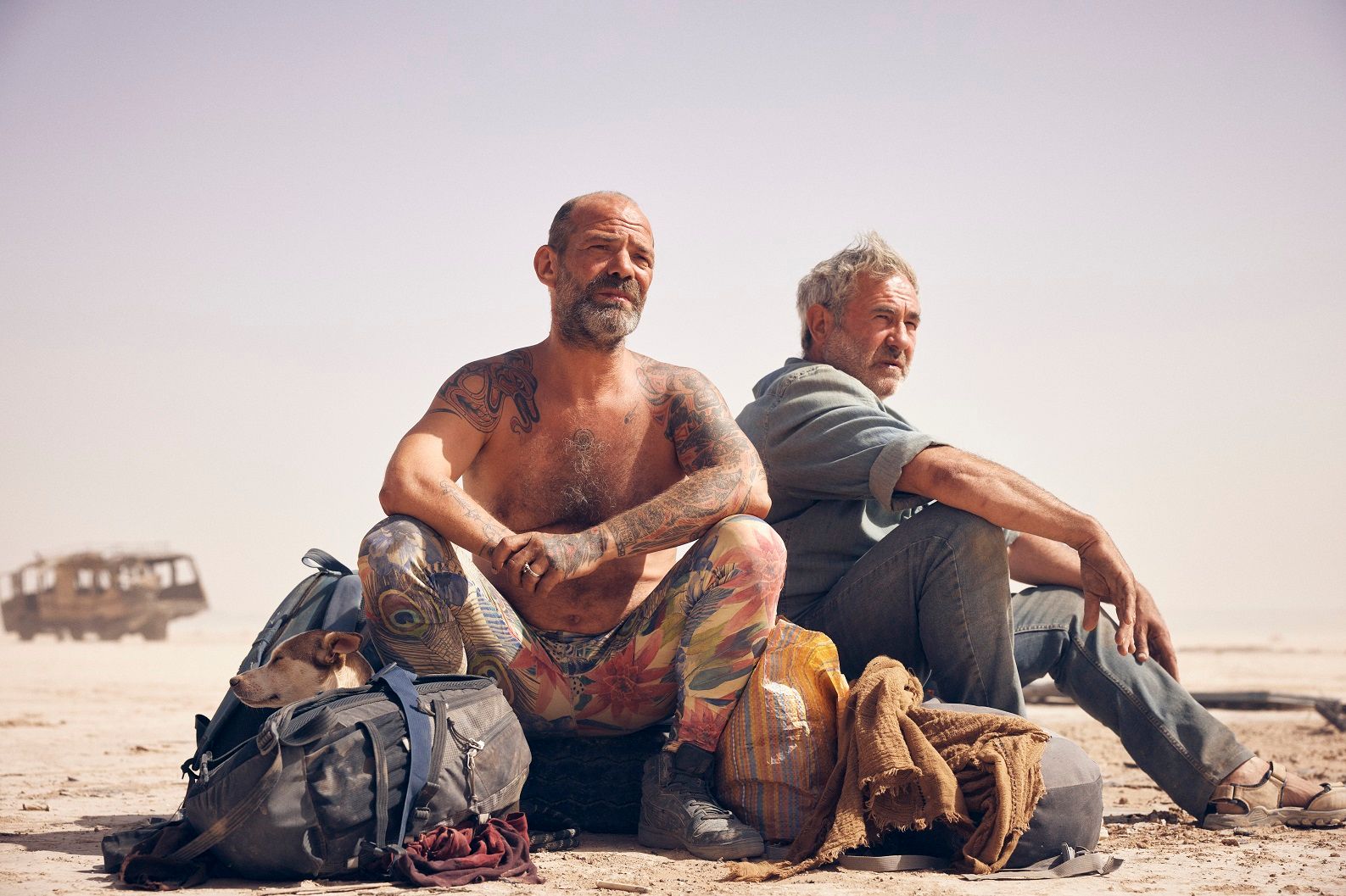 Two men sit on a dry, sandy plain next to several large bags, with a dog resting its head on one of the packs. The man in the foreground is shirtless with extensive tattoos, while a rusted, abandoned bus sits in the distant, hazy background.