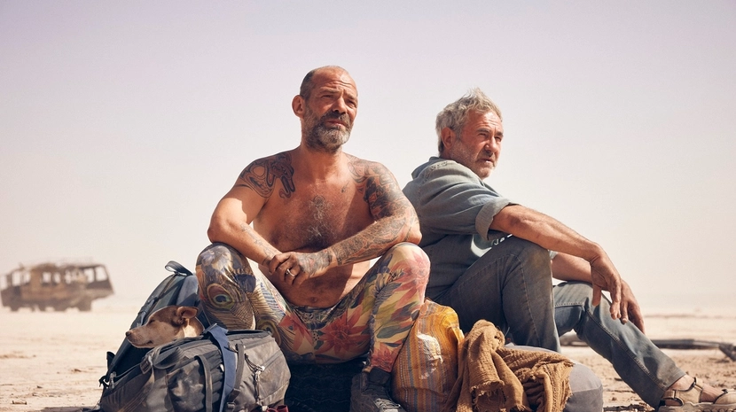 Two men sit on a dry, sandy plain next to several large bags, with a dog resting its head on one of the packs. The man in the foreground is shirtless with extensive tattoos, while a rusted, abandoned bus sits in the distant, hazy background.