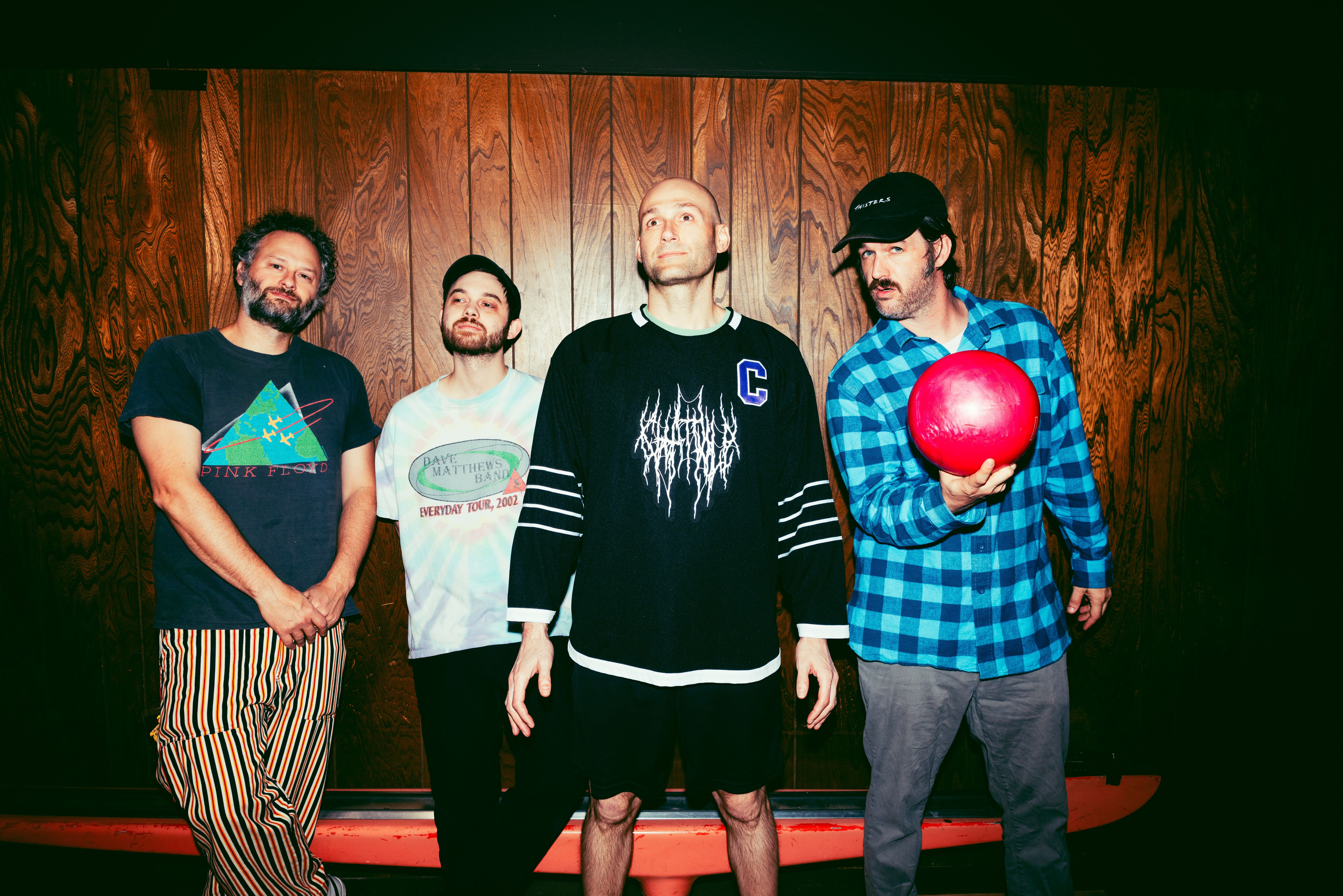 Four men pose against a wood-paneled wall, including one holding a bright red bowling ball. They are dressed in casual, eclectic clothing, and the lighting is sharp and high-contrast.
