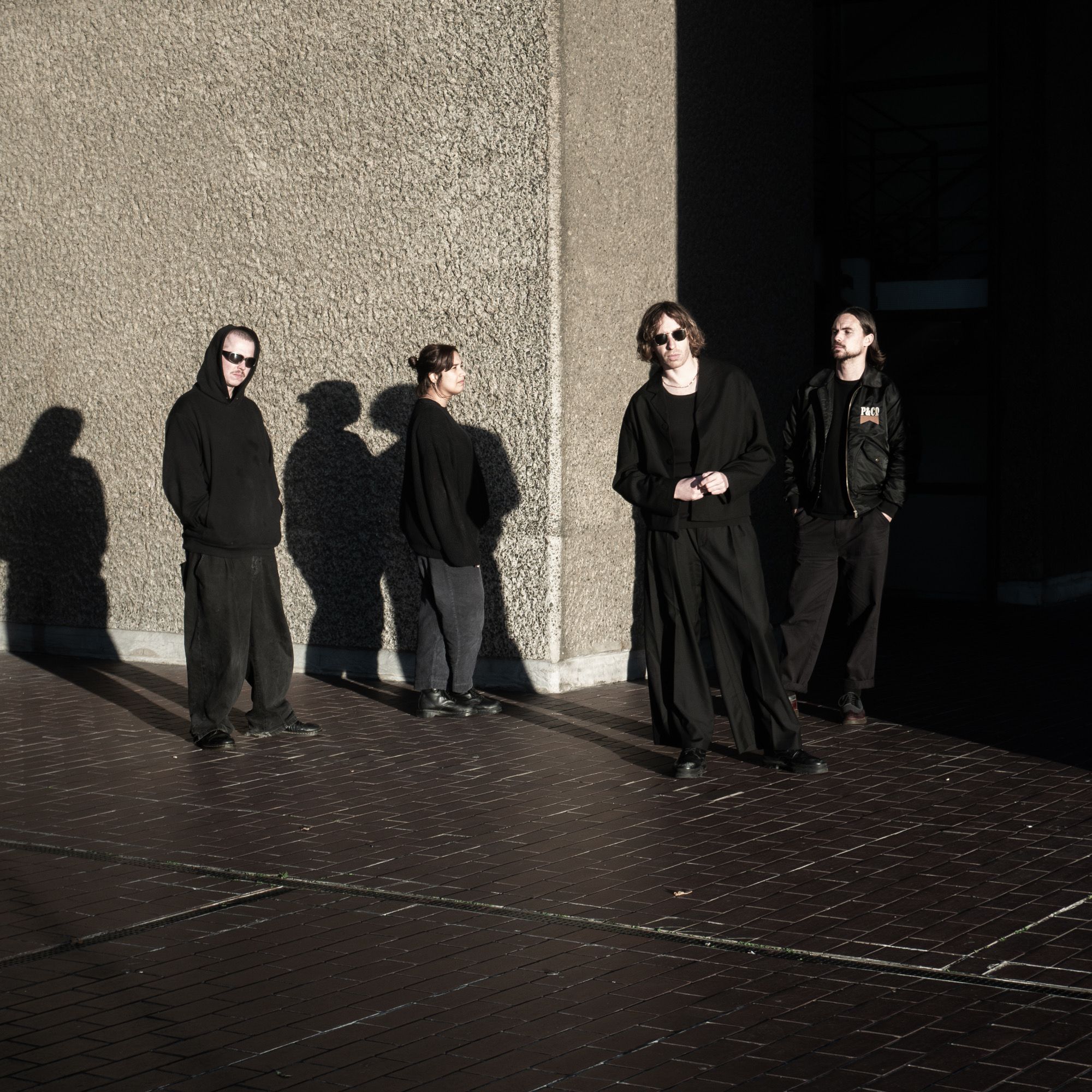 Four people dressed in dark, minimalist streetwear stand in a courtyard against a textured concrete wall under bright, high-contrast sunlight. Long, sharp shadows stretch across the brick ground as the group poses with a somber, cinematic aesthetic.