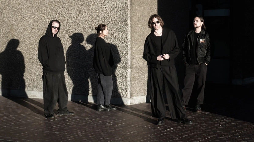 Four people dressed in dark, minimalist streetwear stand in a courtyard against a textured concrete wall under bright, high-contrast sunlight. Long, sharp shadows stretch across the brick ground as the group poses with a somber, cinematic aesthetic.
